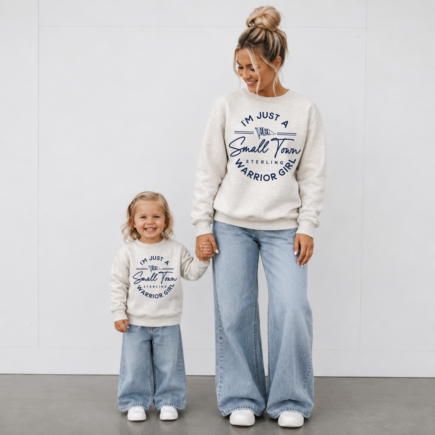 Woman and toddler wearing matching natural heather I'm Just a Small Town Warrior Girl Sterling football crewneck sweatshirts with light wash jeans for game day.