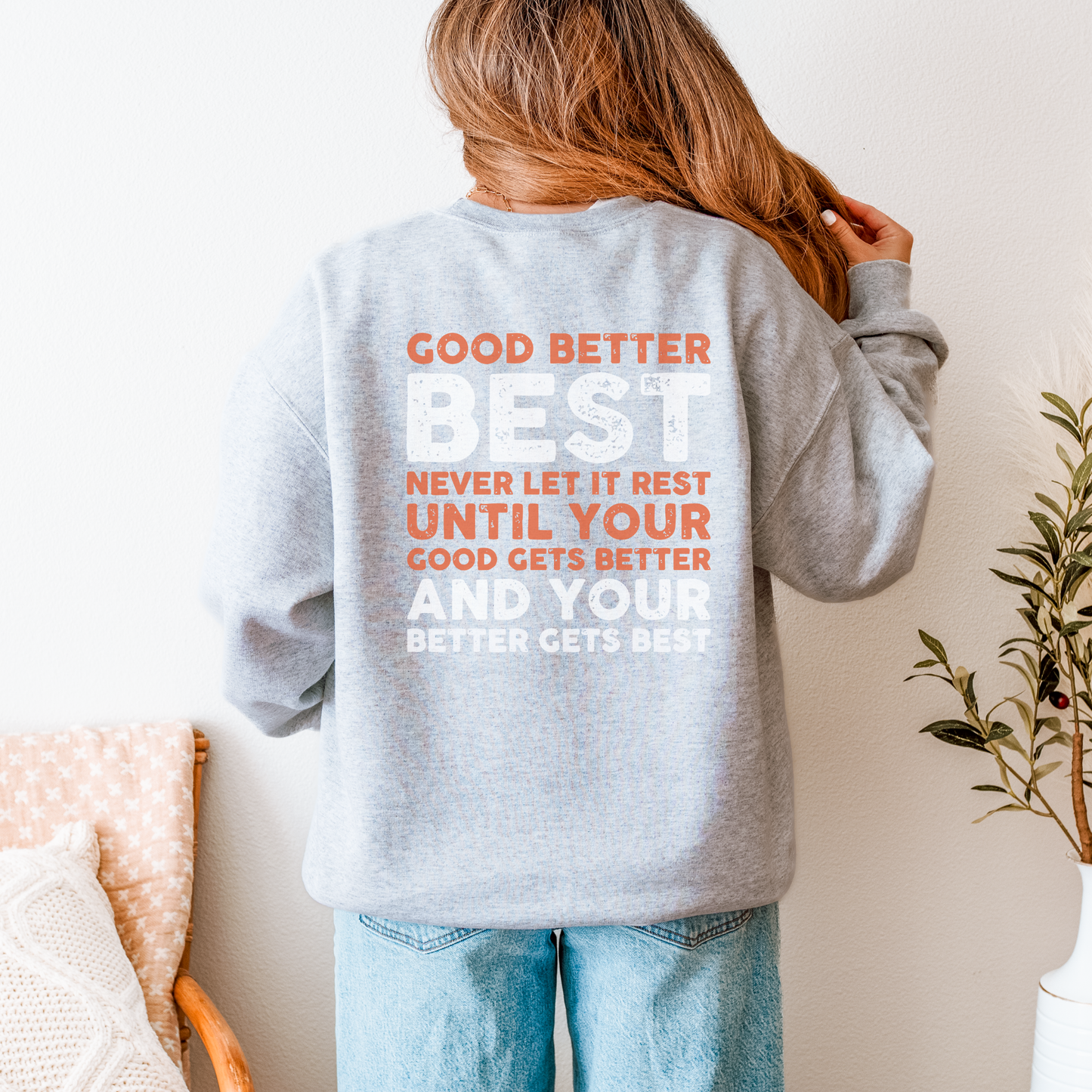 Person wearing a gray sweatshirt with motivational text on the back, standing in a room with a plant and chair.