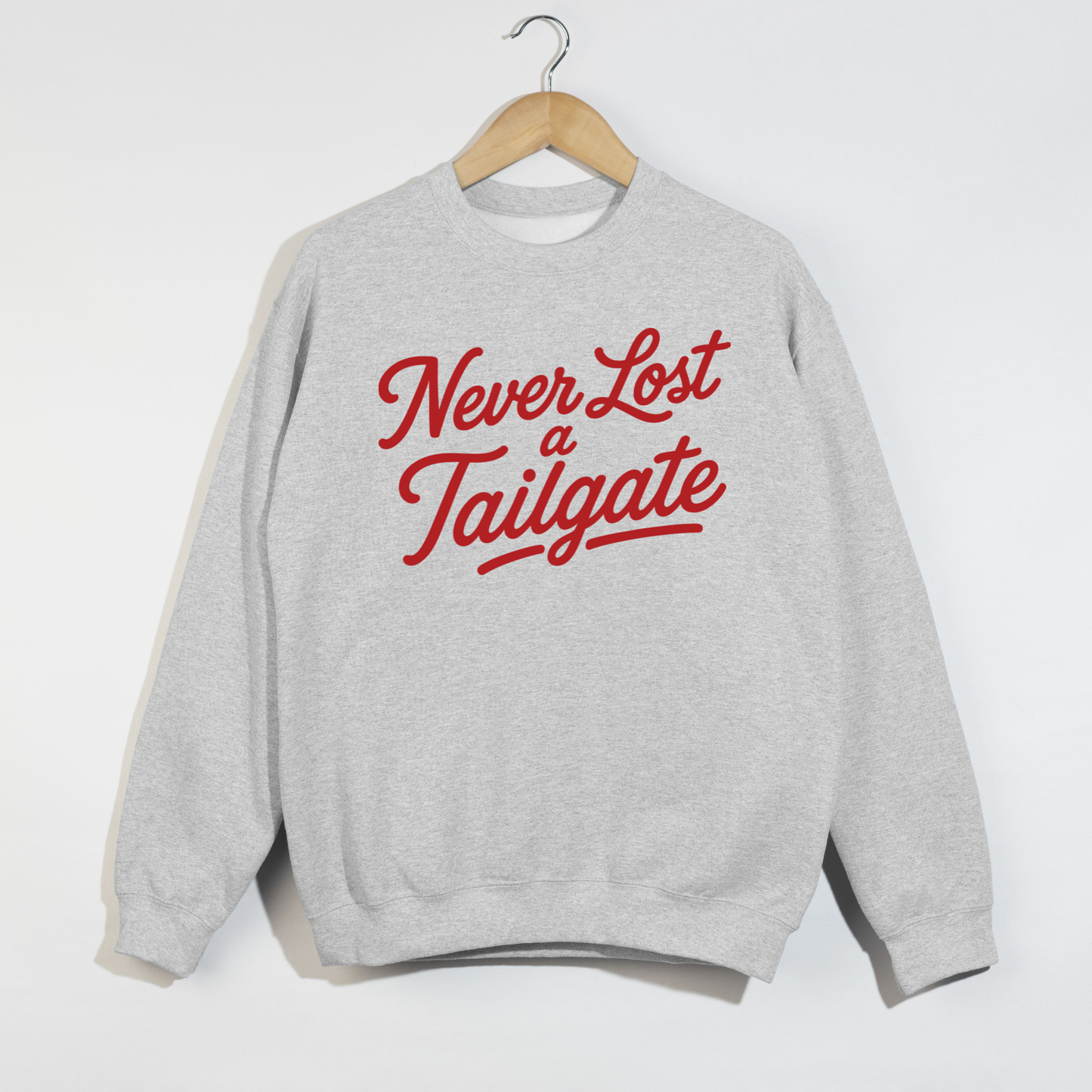 Ash gray sweatshirt featuring the phrase "Never Lost a Tailgate" in bold crimson script lettering hanging on a wooden hanger.
