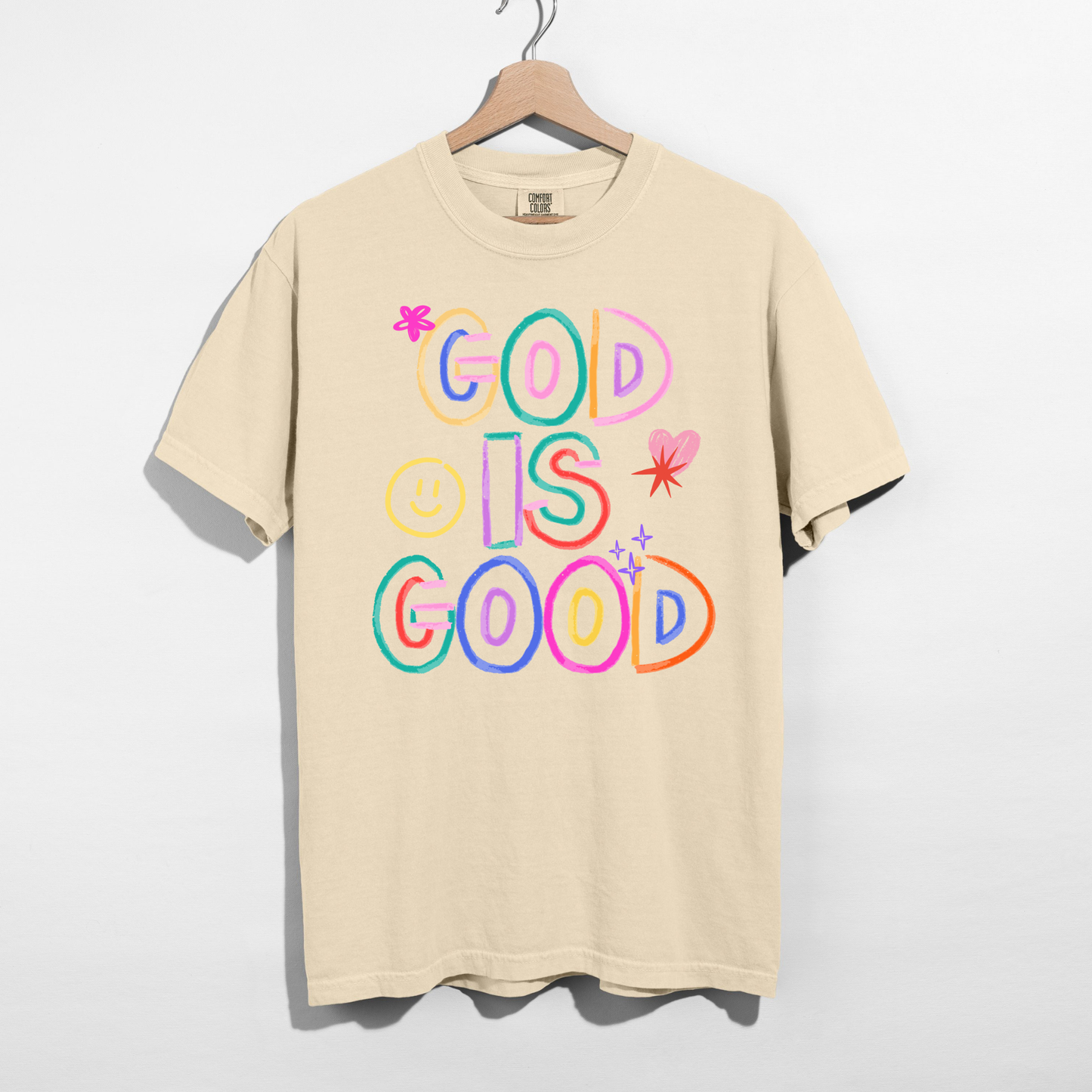 Beige t-shirt with 'God is Good' text in colorful letters on a hanger against a white background