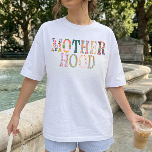 Woman wearing oversized white motherhood graphic tee with colorful patchwork lettering near outdoor fountain while holding iced coffee.