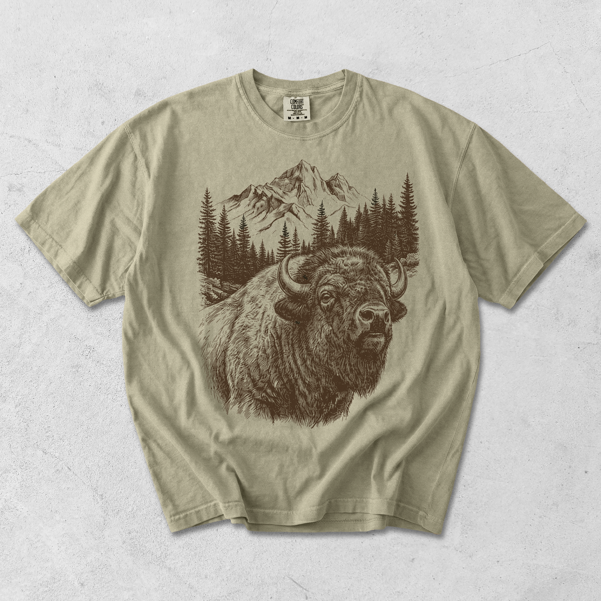 Khaki buffalo mountain graphic tee flat lay featuring detailed wildlife illustration with evergreen forest and mountain scenery.
