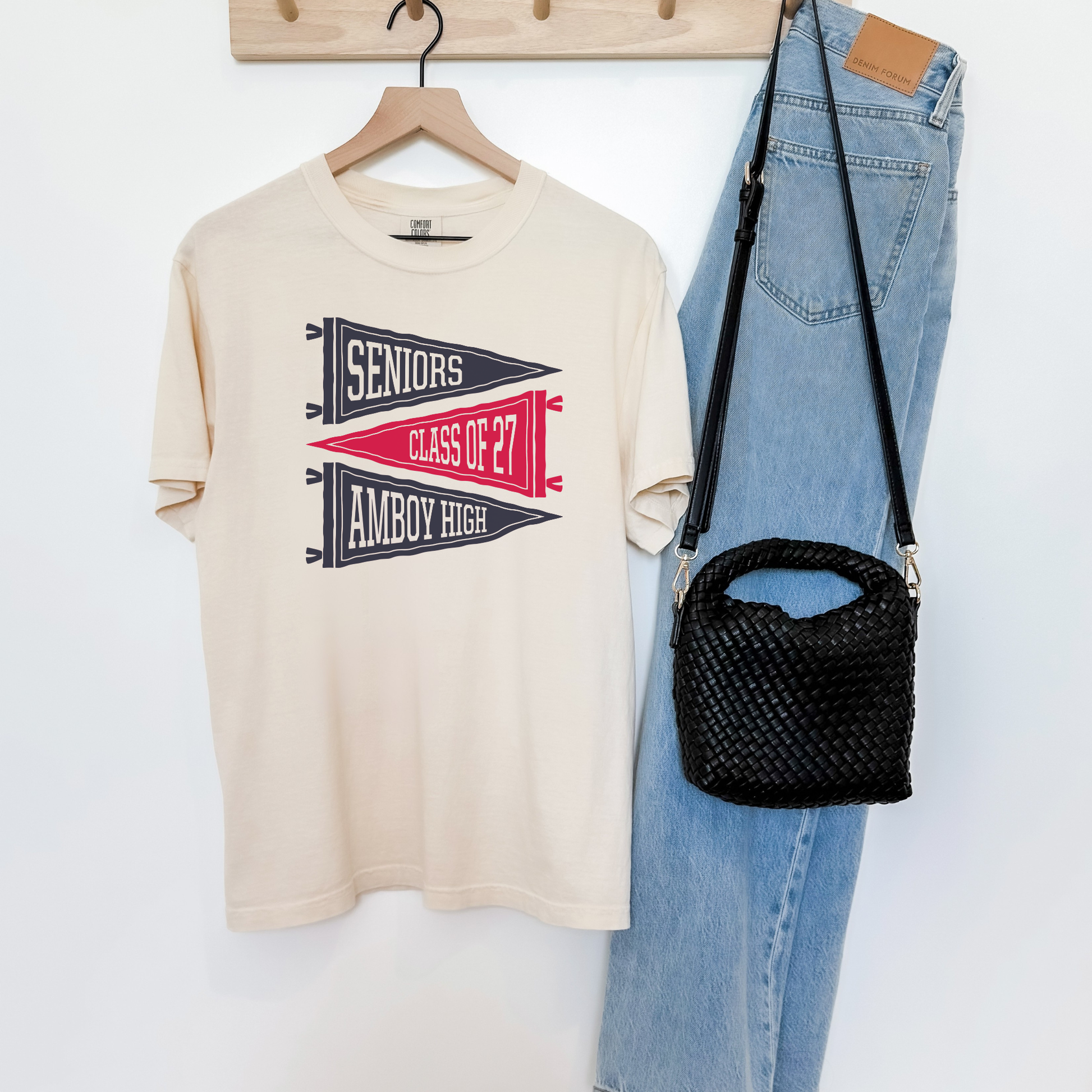 Ivory Amboy High School Class of 2027 pennant graphic tee hanging beside light wash denim jeans and a black woven handbag.