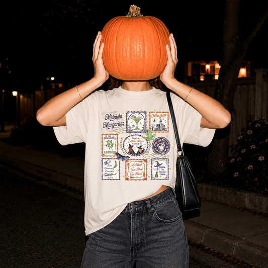 Woman wearing an ivory Midnight Margaritas vintage Halloween graphic tee with witchy postage stamp collage, holding a pumpkin over her face and styling the shirt with dark denim jeans.