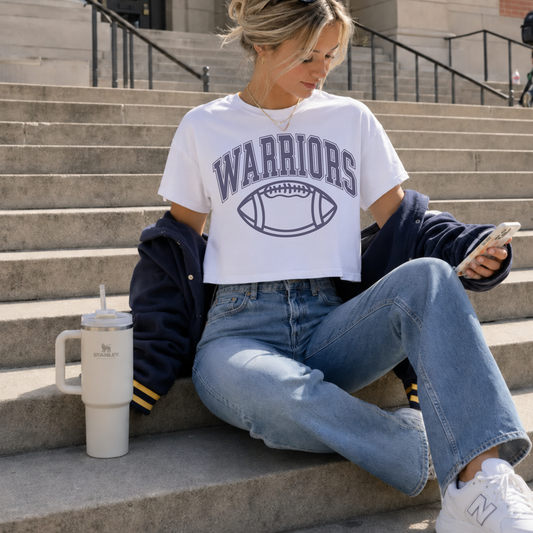 Sterling Warriors football graphic boxy crop tee styled with light wash jeans and navy varsity jacket on stadium steps.