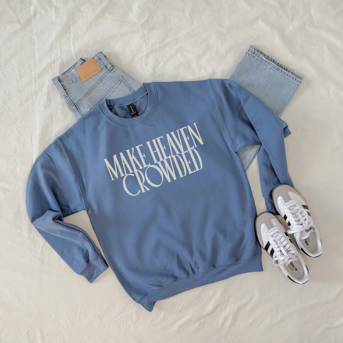 Blue sweatshirt with 'MAKE HEAVEN CROWDED' text, jeans, and sneakers on a light background
