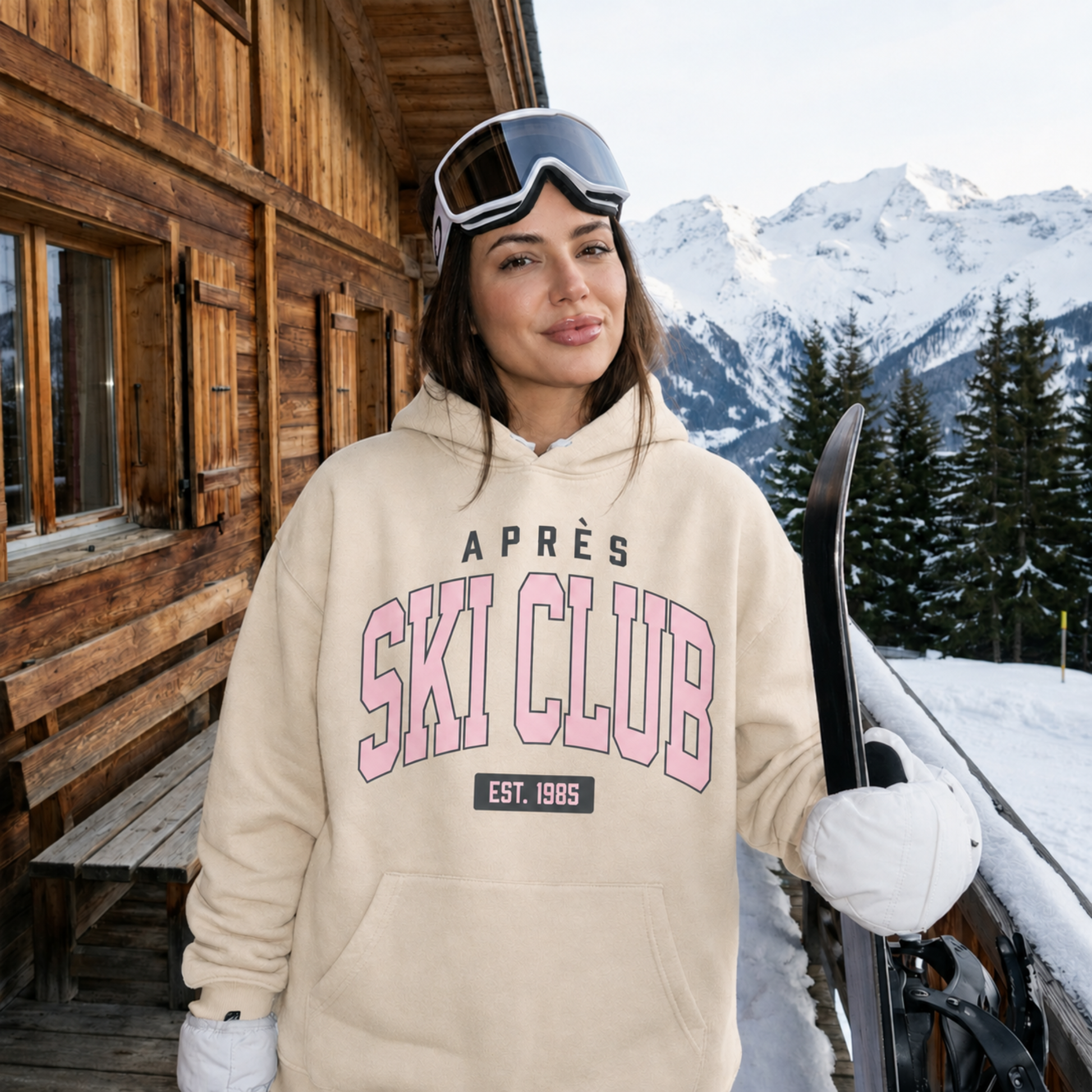 Woman wearing a Sand Après Ski Club hoodie holding skis outside a cozy mountain cabin.