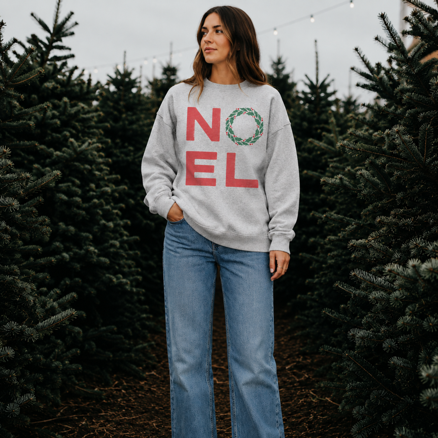 Woman wearing an oatmeal heather NOEL Christmas sweatshirt at a Christmas tree farm.