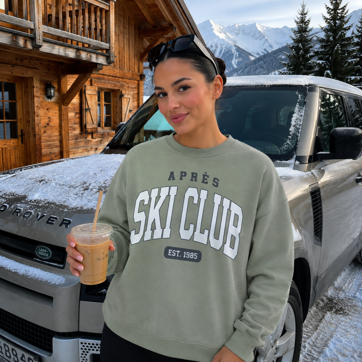 Woman wearing a sage green Après Ski Club sweatshirt while holding iced coffee in front of a snowy mountain cabin and SUV.