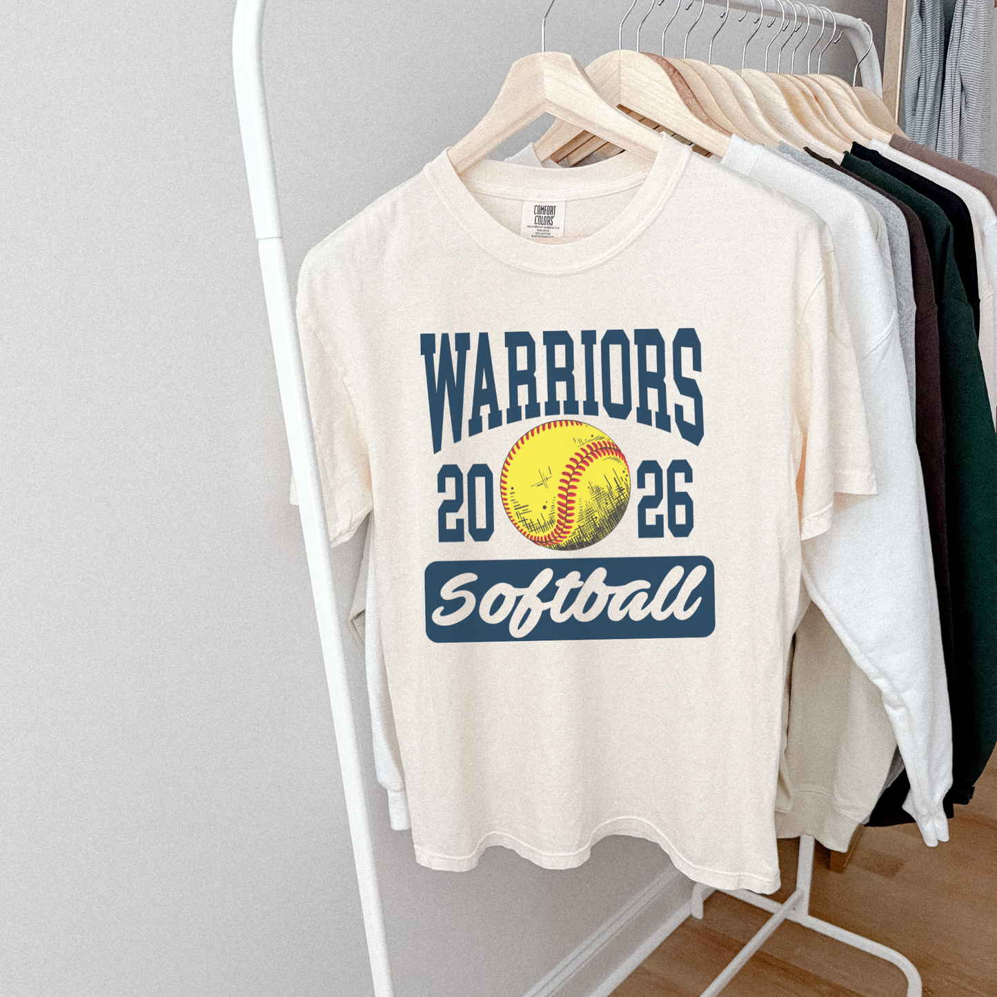 White t-shirt with 'Warriors 2026 Softball' design on a rack against a gray wall.