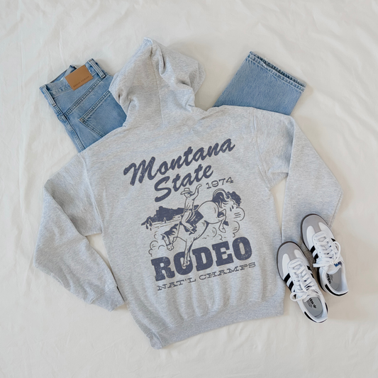 Gray hoodie with 'Montana State Rodeo' design, jeans, and sneakers on a light background