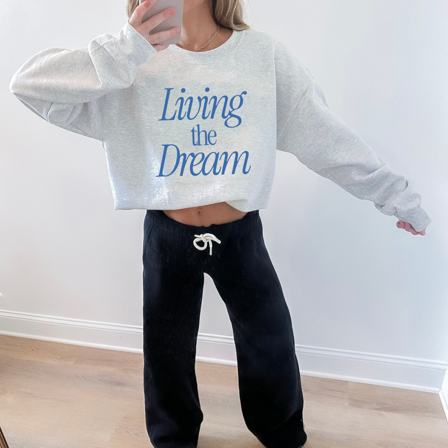 Person wearing a gray sweatshirt with 'Living the Dream' text and black pants, standing against a white wall.