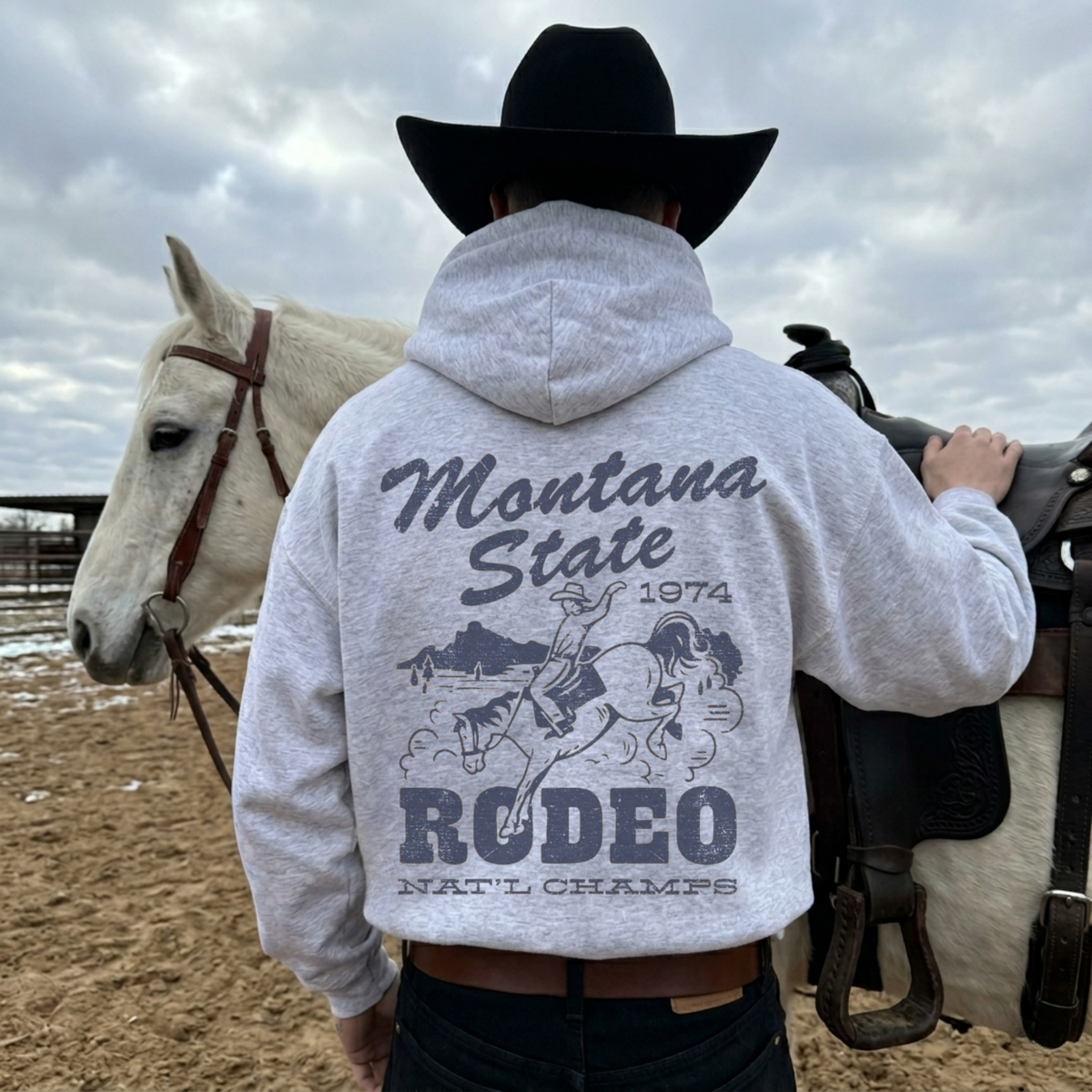 Back view of ash grey Montana State Rodeo western hoodie featuring distressed cowboy rodeo graphic beside white horse.