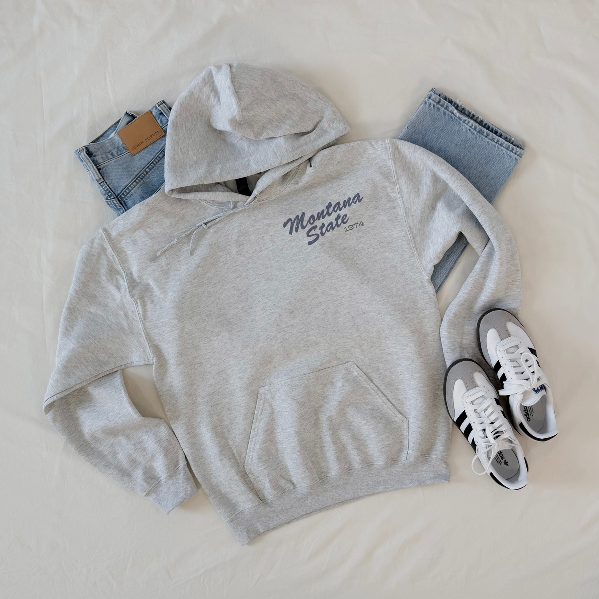 Ash grey western hoodie with small distressed Montana State 1974 left chest graphic styled with denim and sneakers.