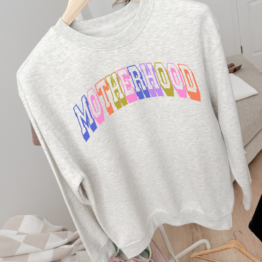 Ash grey motherhood graphic crewneck sweatshirt with colorful retro collegiate lettering hanging in cozy neutral room.