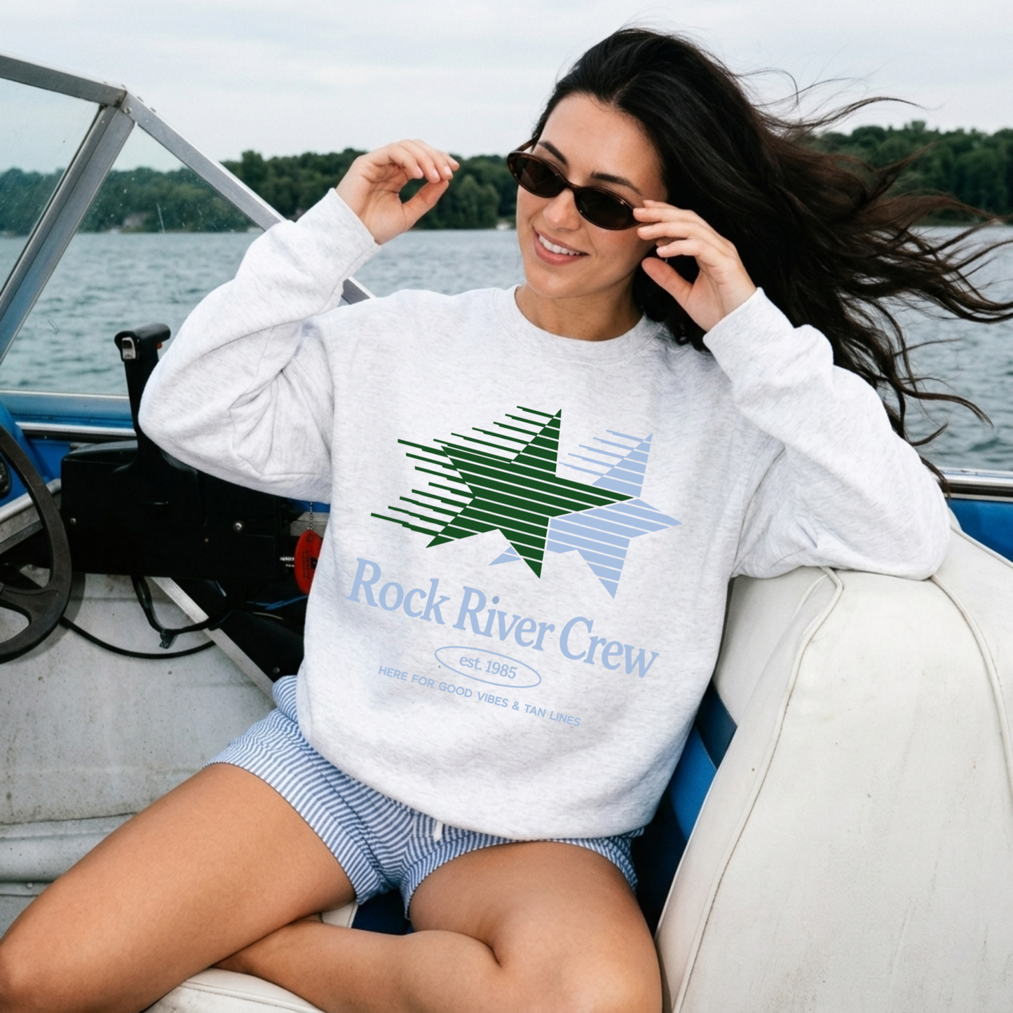 Woman wearing a 'Rock River Crew' sweatshirt on a boat by a lake.
