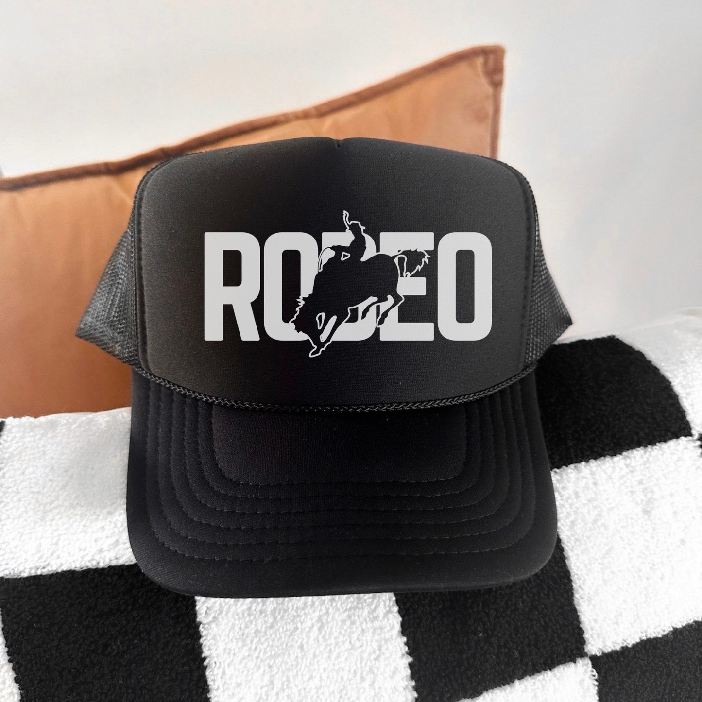 Black trucker hat with 'RODEO' and bull graphic on a checkered blanket with brown pillow
