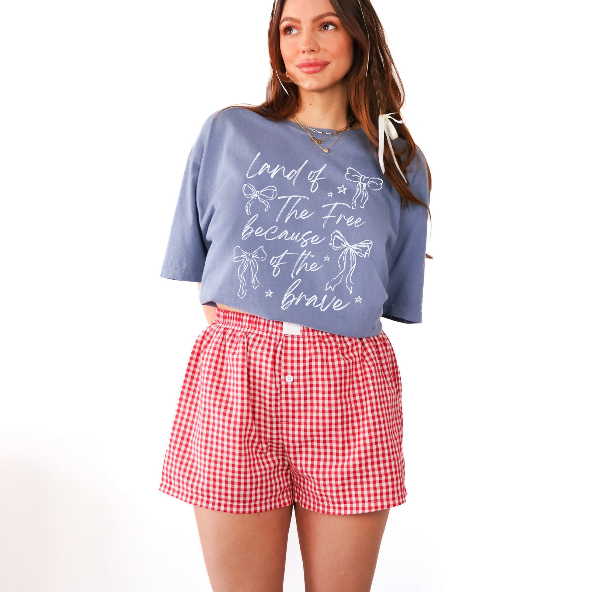 A woman wearing a blue graphic t-shirt with the text 'Land of the Free Because of the Brave' and red decorative bows printed on it, paired with red and white checkered shorts.