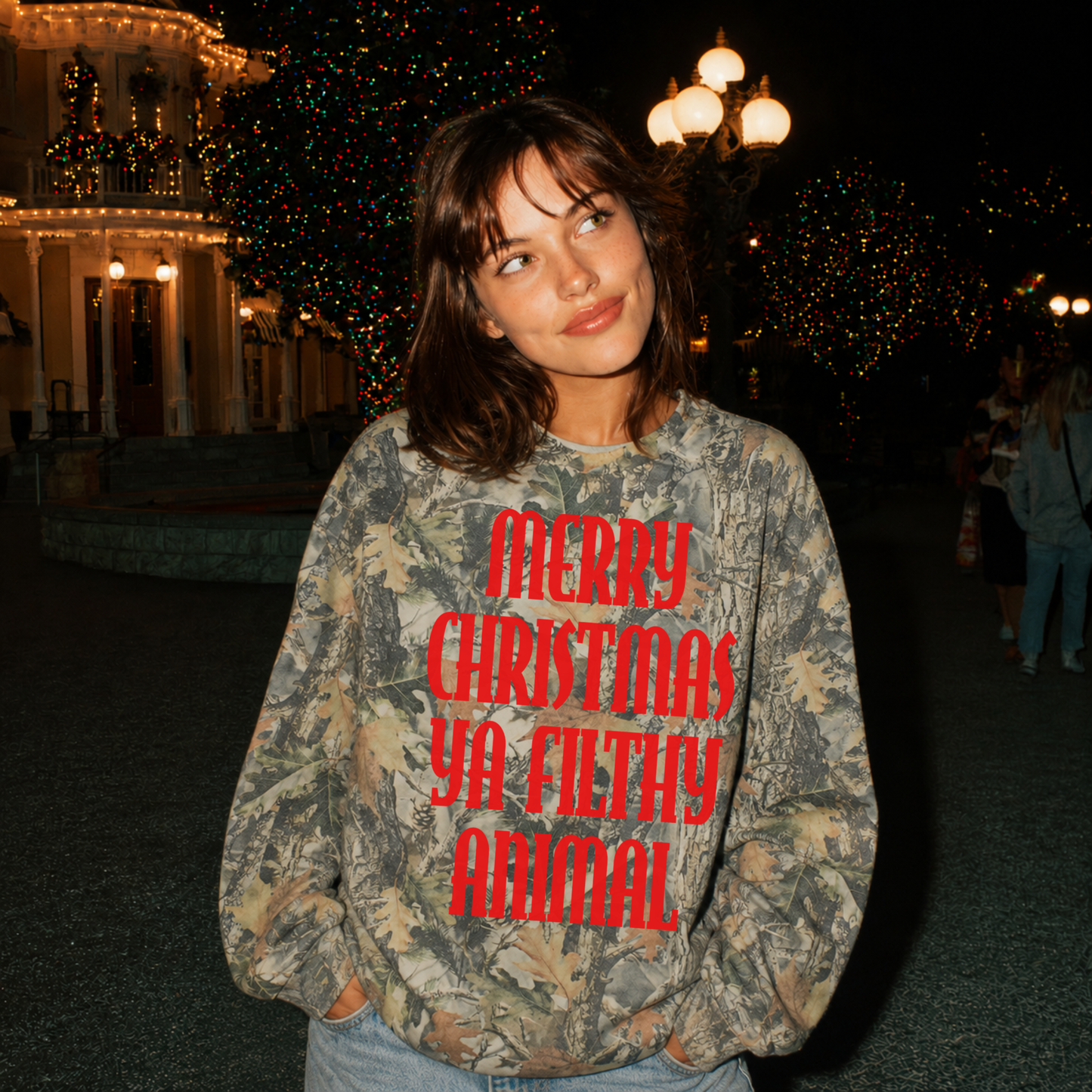 Woman wearing a premium oversized woodland camo Merry Christmas Ya Filthy Animal sweatshirt at a festive Christmas lights display.