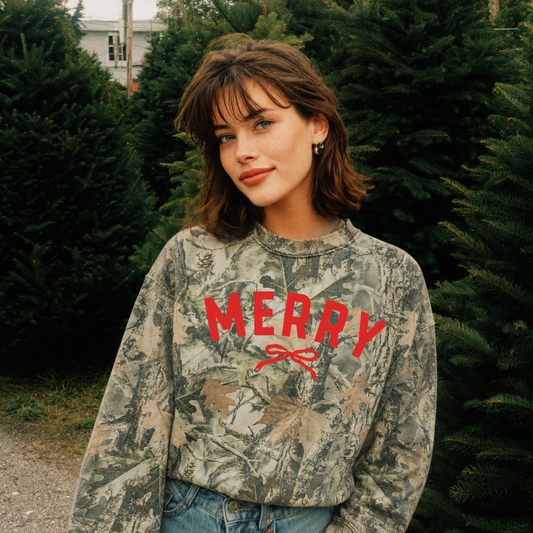 Woman wearing a premium oversized camo Merry Christmas crewneck with red bow graphic at a Christmas tree farm.