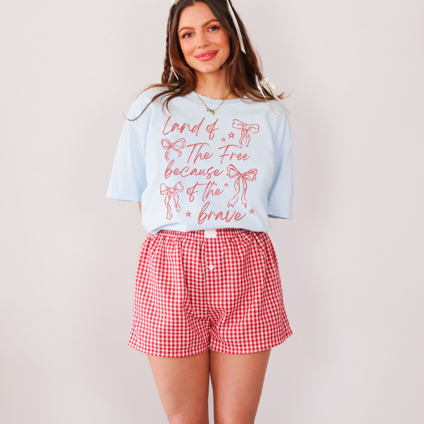 A woman wearing a chambray graphic t-shirt with the text 'Land of the Free Because of the Brave' and red decorative bows printed on it, paired with red and white checkered shorts.