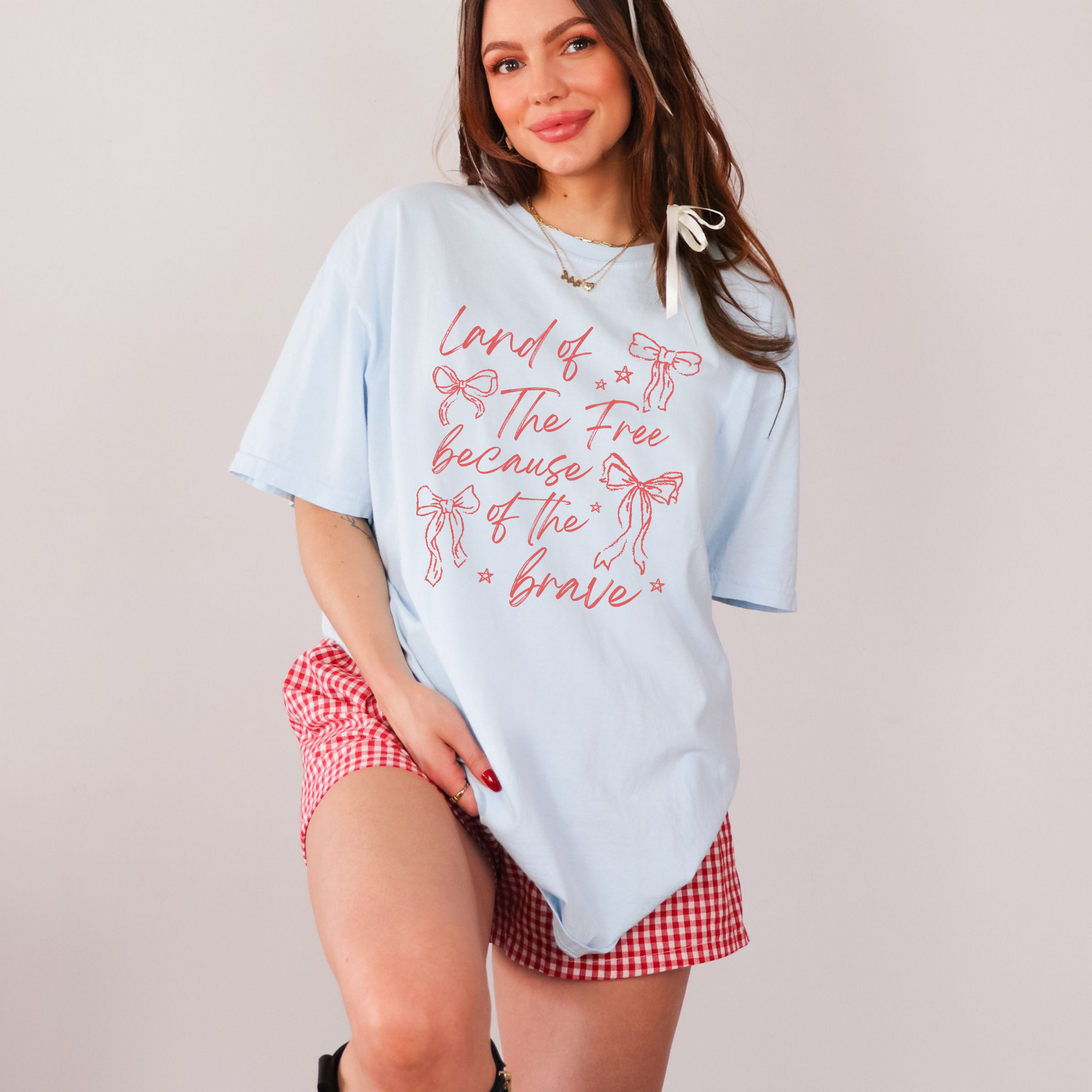 Woman wearing a chambray Comfort Colors 1717 graphic tee with pink “Land of the Free Because of the Brave” patriotic bow design while holding a red, white, and blue popsicle on a lakeside dock.