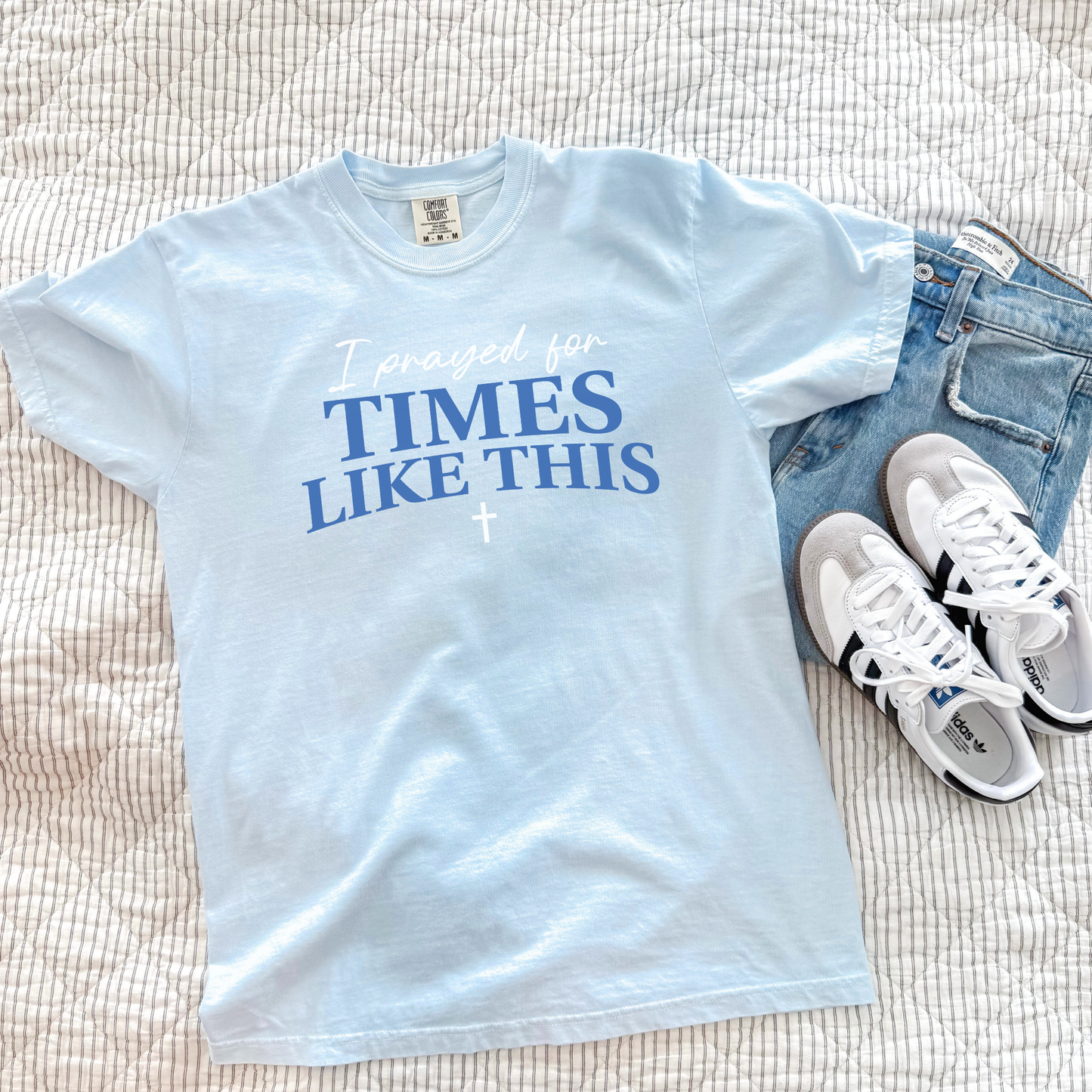 Light blue t-shirt with text 'I prayed for times like this' on a bed with jeans and sneakers.