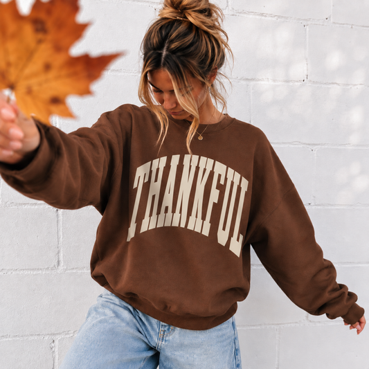 Woman wearing chestnut brown Thankful sweatshirt with cream varsity lettering and jeans for a casual fall outfit