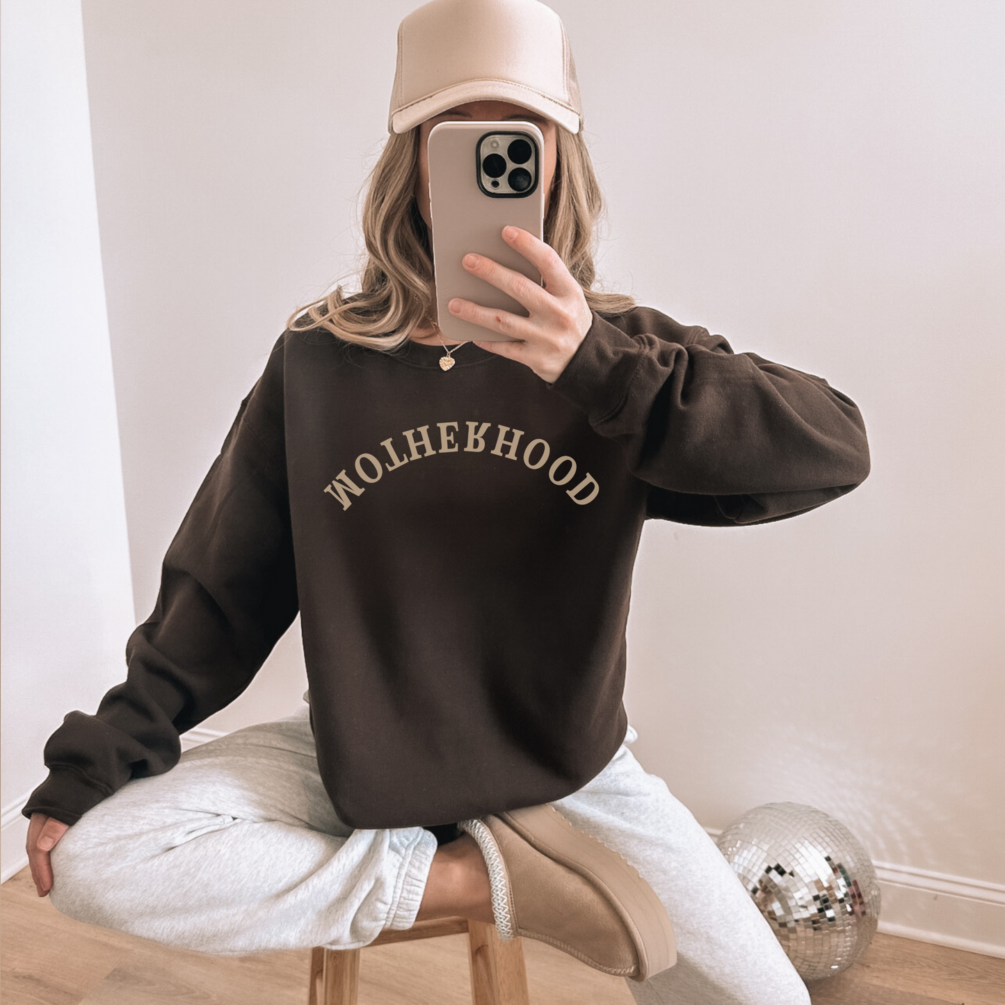 Person wearing a brown sweatshirt with 'Motherhood' text, taking a mirror selfie.