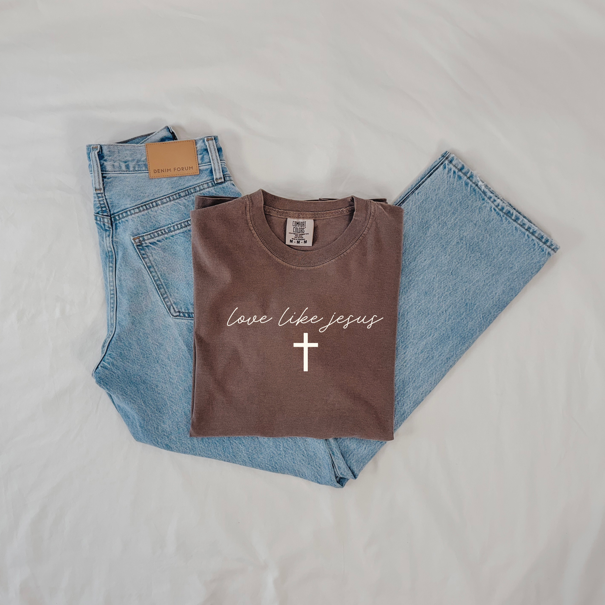 Folded espresso Comfort Colors “Love Like Jesus” Christian tee styled with light wash denim jeans, flat lay.