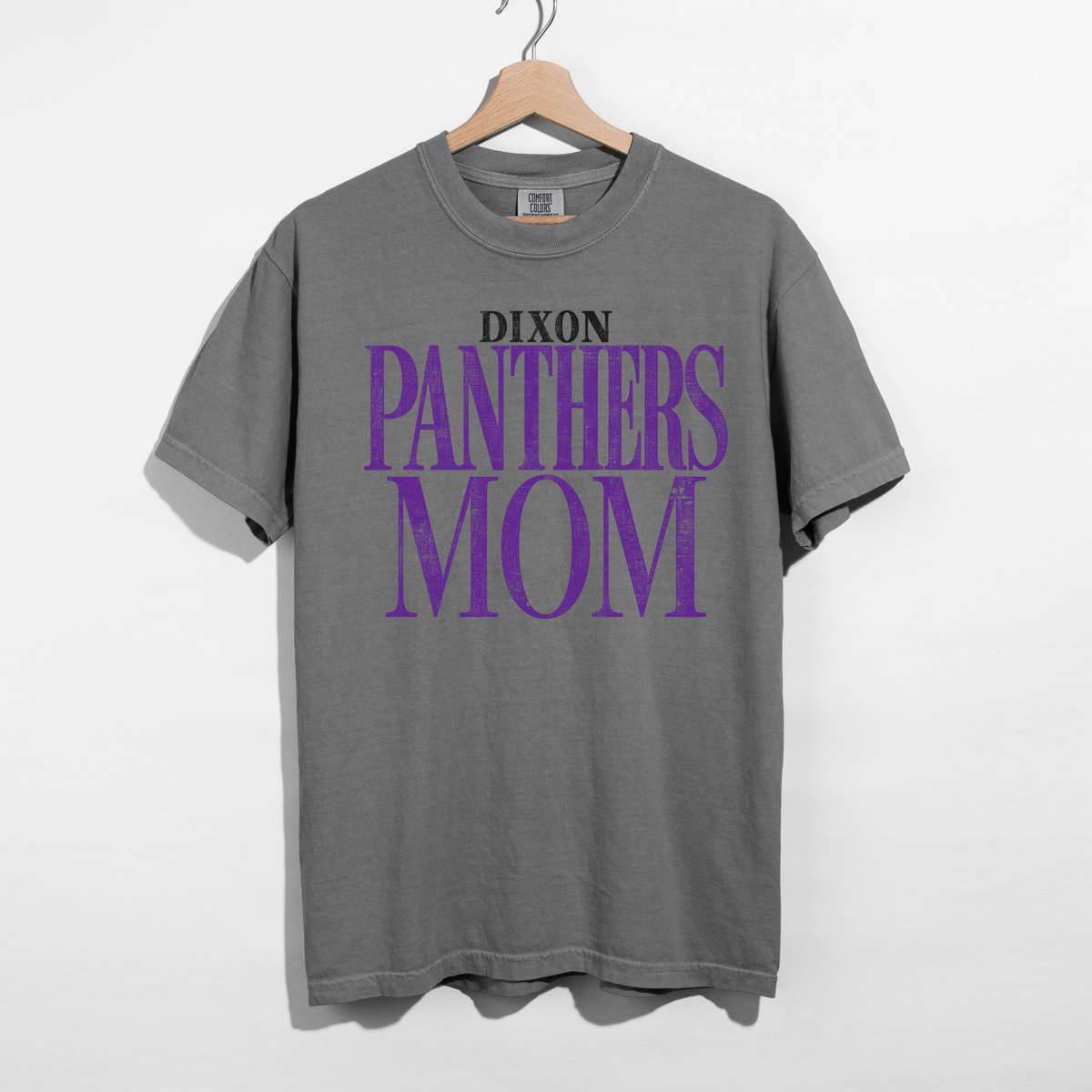 A heather gray short-sleeve t-shirt hanging on a wooden hanger against a white wall. The shirt features a graphic print on the front with the text 'DIXON PANTHERS MOM' in black and purple.