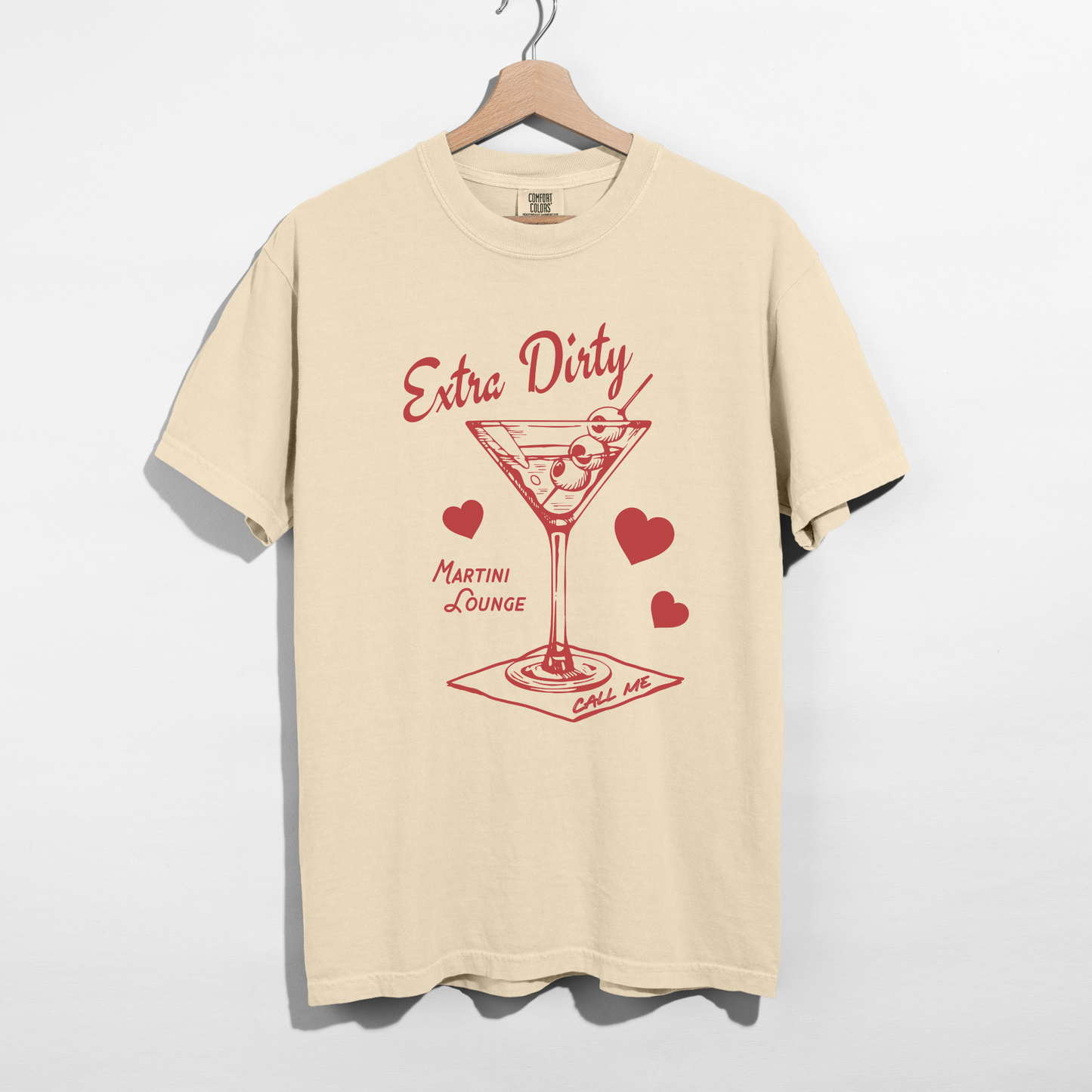 Ivory Comfort Colors Extra Dirty Martini graphic tee on wooden hanger against white background.