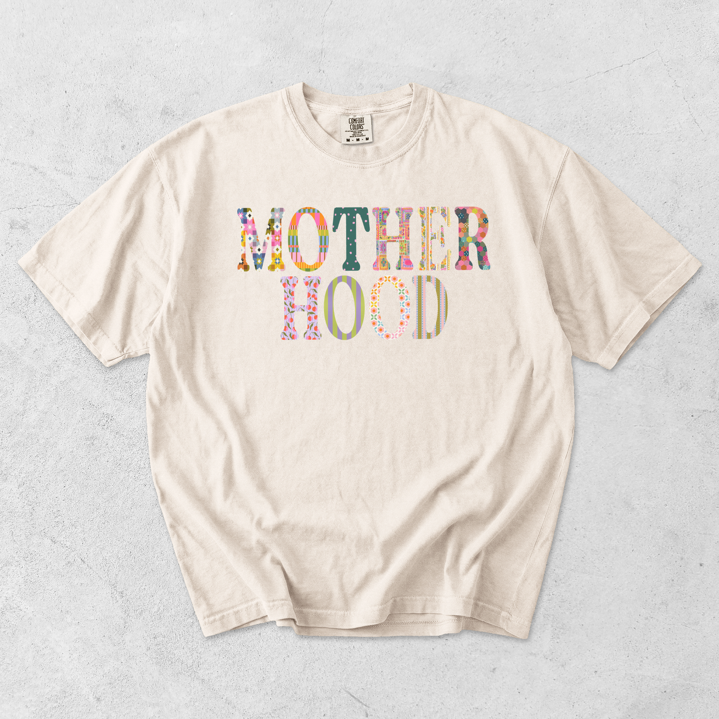 Ivory Comfort Colors 1717 motherhood graphic tee with colorful mixed-pattern patchwork lettering on light textured background.