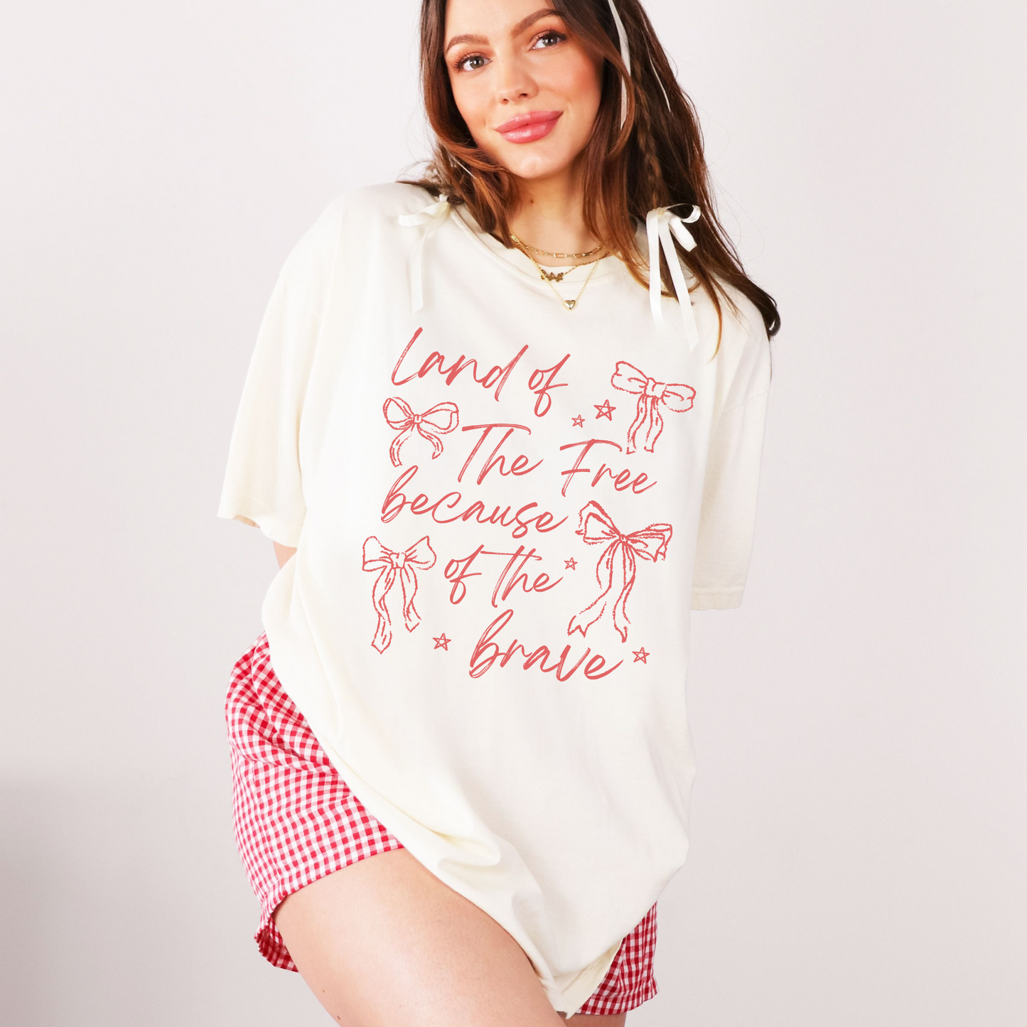 A woman wearing a white graphic t-shirt with the text 'Land of the Free Because of the Brave' and red decorative bows printed on it, paired with red and white checkered shorts.