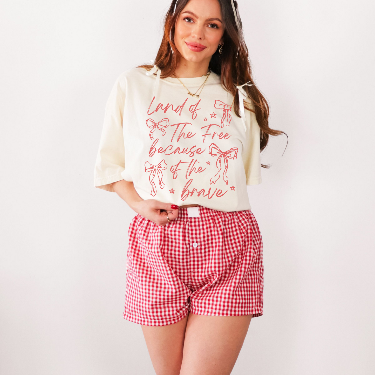 A woman wearing a ivory graphic t-shirt with the text 'Land of the Free Because of the Brave' and red decorative bows printed on it, paired with red and white checkered shorts.