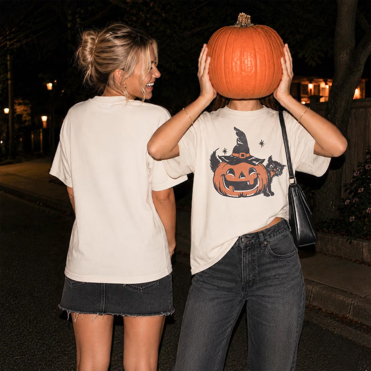 Two women styling casual Halloween outfits at night, one wearing an ivory pumpkin and black cat graphic tee with dark denim jeans and holding a pumpkin.