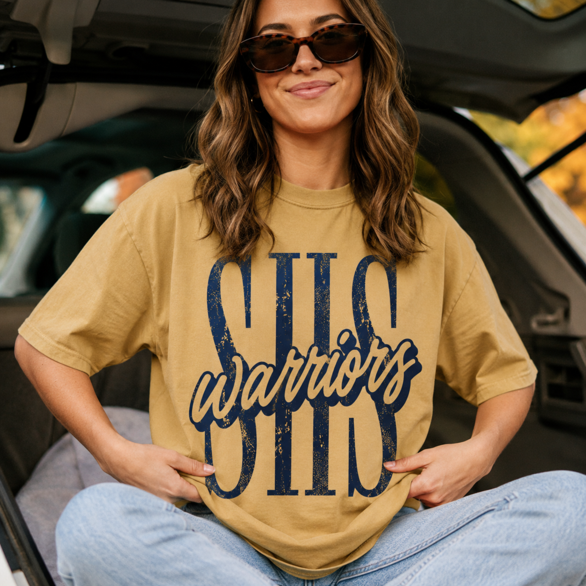 Woman wearing a mustard Sterling Warriors SHS graphic t-shirt with oversized navy distressed lettering and script Warriors while sitting in the back of an SUV for a casual game day look.