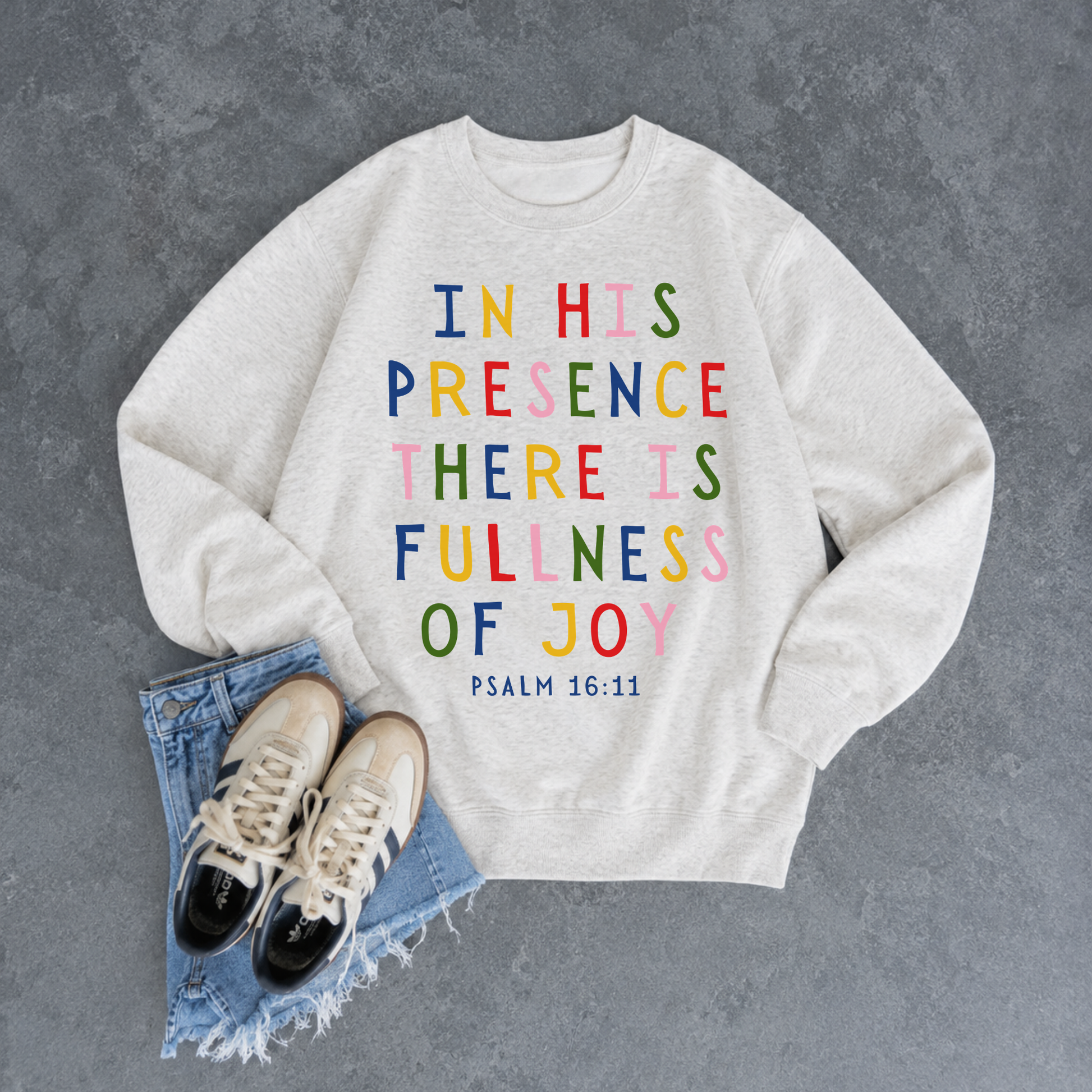 Oatmeal heather Christian crewneck sweatshirt featuring the colorful Bible verse "In His Presence There Is Fullness of Joy" from Psalm 16:11 styled with denim shorts and sneakers.