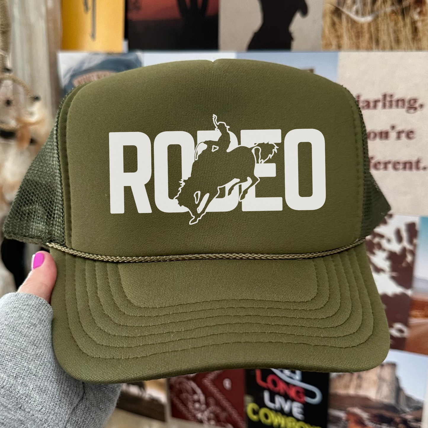 Green cap with 'RODEO' and horse graphic held by a hand against a blurred background