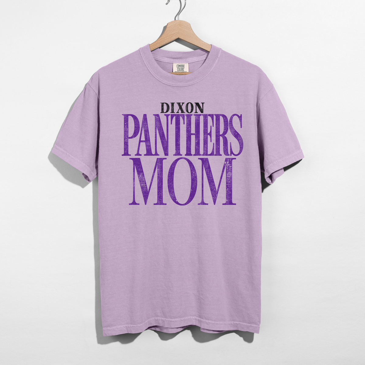 A light purple short-sleeve t-shirt hanging on a wooden hanger against a white wall. The shirt features a graphic print on the front reading 'DIXON PANTHERS MOM' in purple and black text.