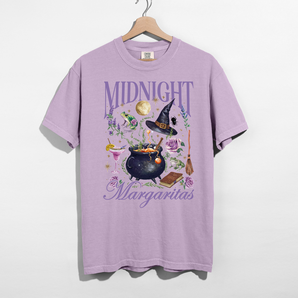 Orchid purple Midnight Margaritas Halloween graphic tee hanging on a wooden hanger, featuring a witch hat, bubbling cauldron, margarita glass, moon, frogs, and lavender floral artwork.