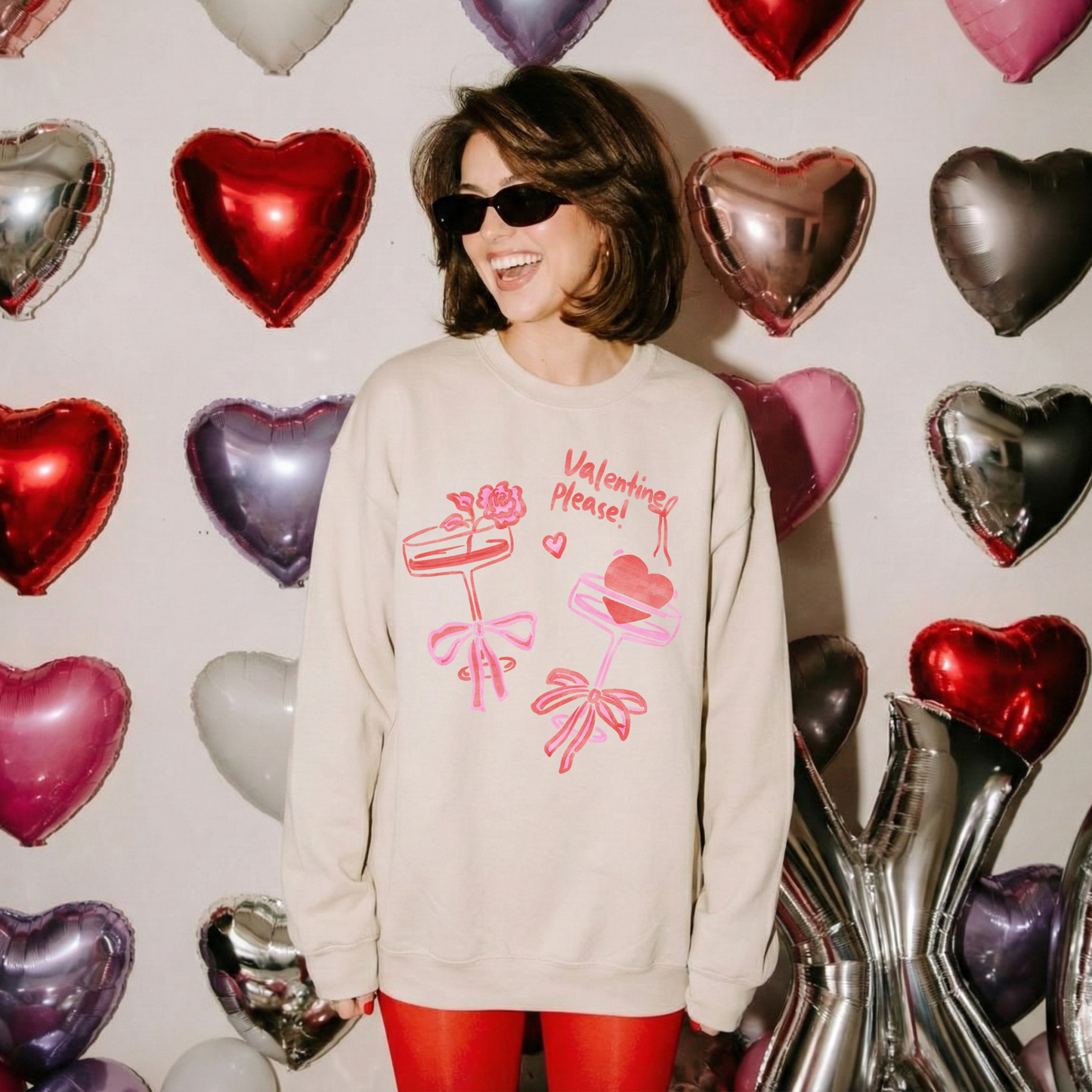 Woman wearing a sand-colored Valentine’s crewneck with pink and red bow designs, smiling in front of metallic heart balloons.