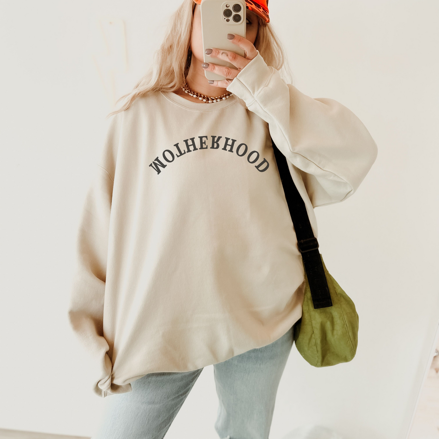 A person wearing a beige crewneck sweatshirt with the text 'Motherhood' on the front, paired with blue jeans and a red hat.
