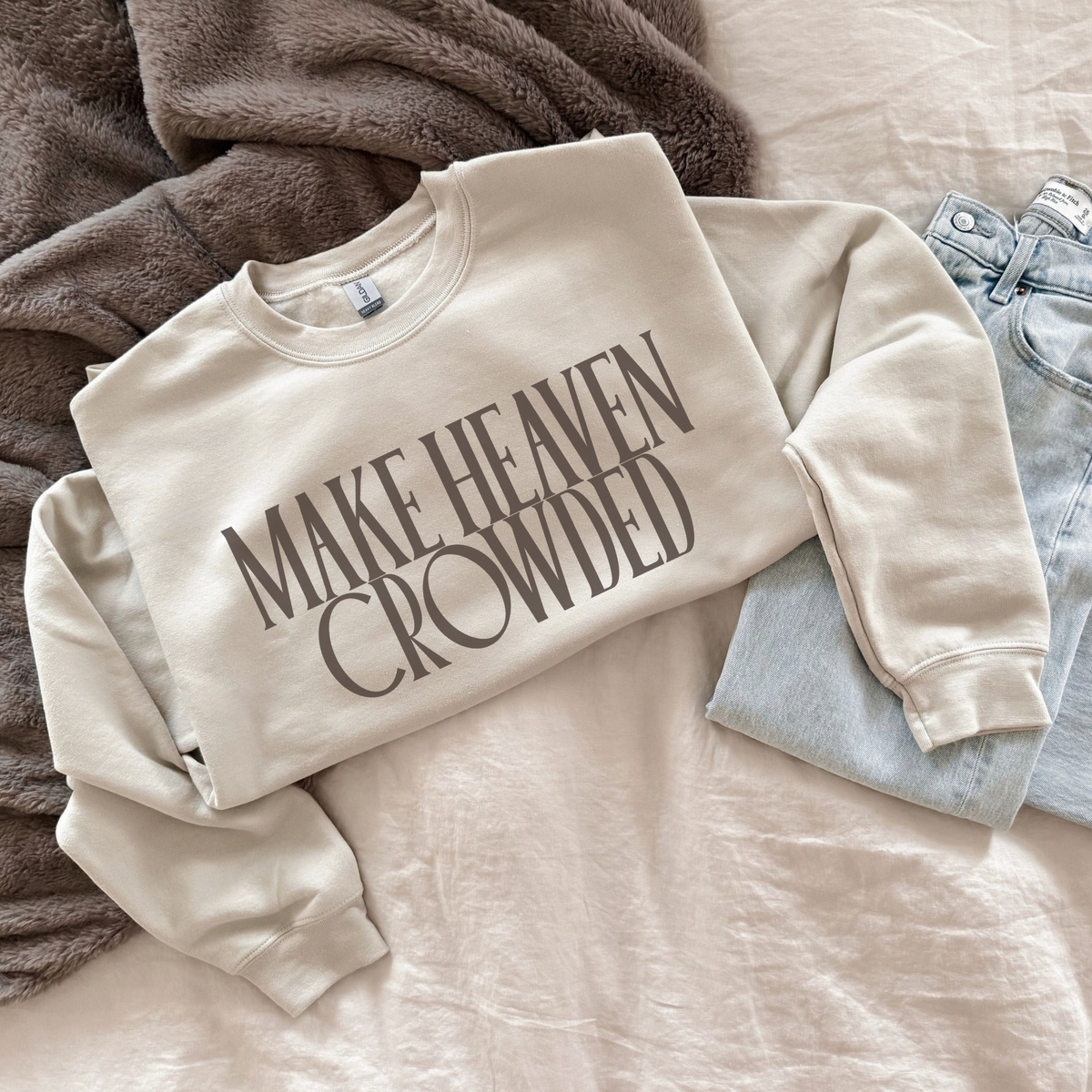 Beige sweatshirt with 'MAKE HEAVEN CROWDED' text on a bed with gray pants and brown blanket.