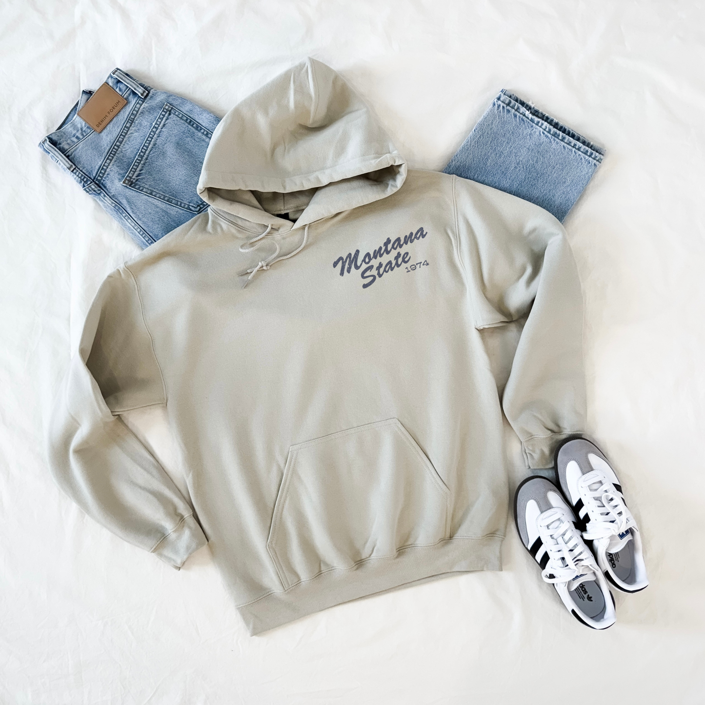 Beige hoodie with 'Montana State' text, blue jeans, and white sneakers on a light background