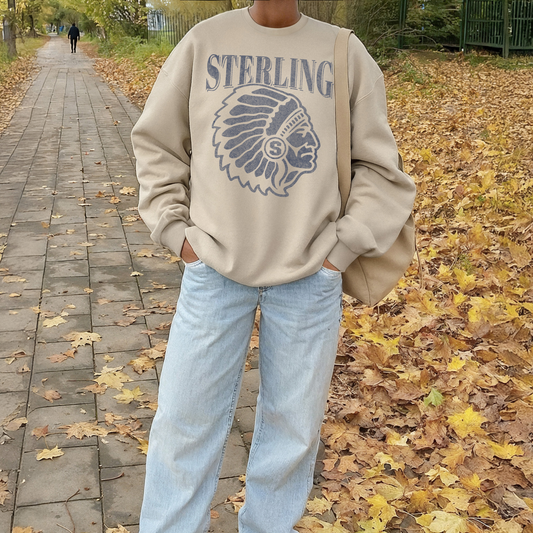 Person wearing a beige sweatshirt with 'STERLING' and a headdress design, standing on a leaf-covered path.