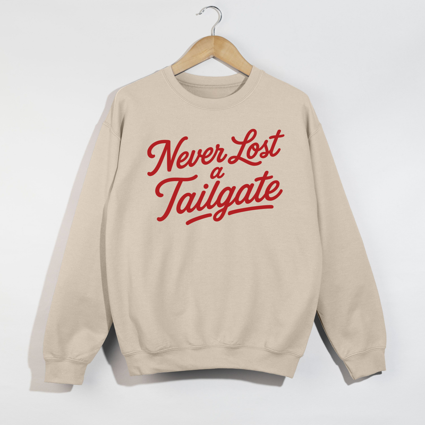 Sand-colored sweatshirt featuring the phrase "Never Lost a Tailgate" in bold crimson script lettering displayed on a wooden hanger.