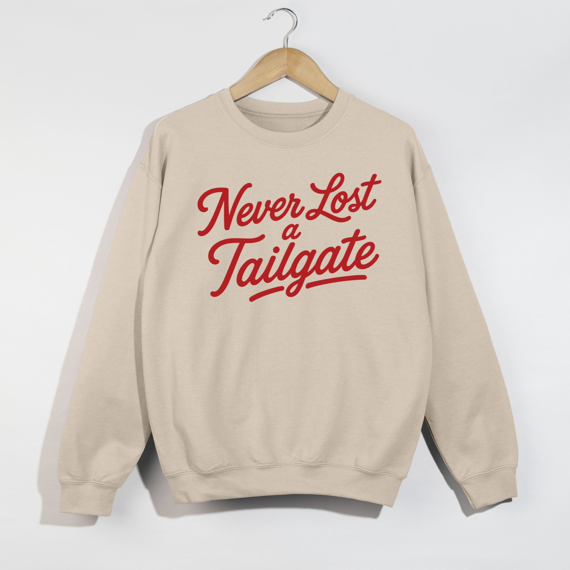 Sand-colored sweatshirt featuring the phrase "Never Lost a Tailgate" in bold crimson script lettering displayed on a wooden hanger.