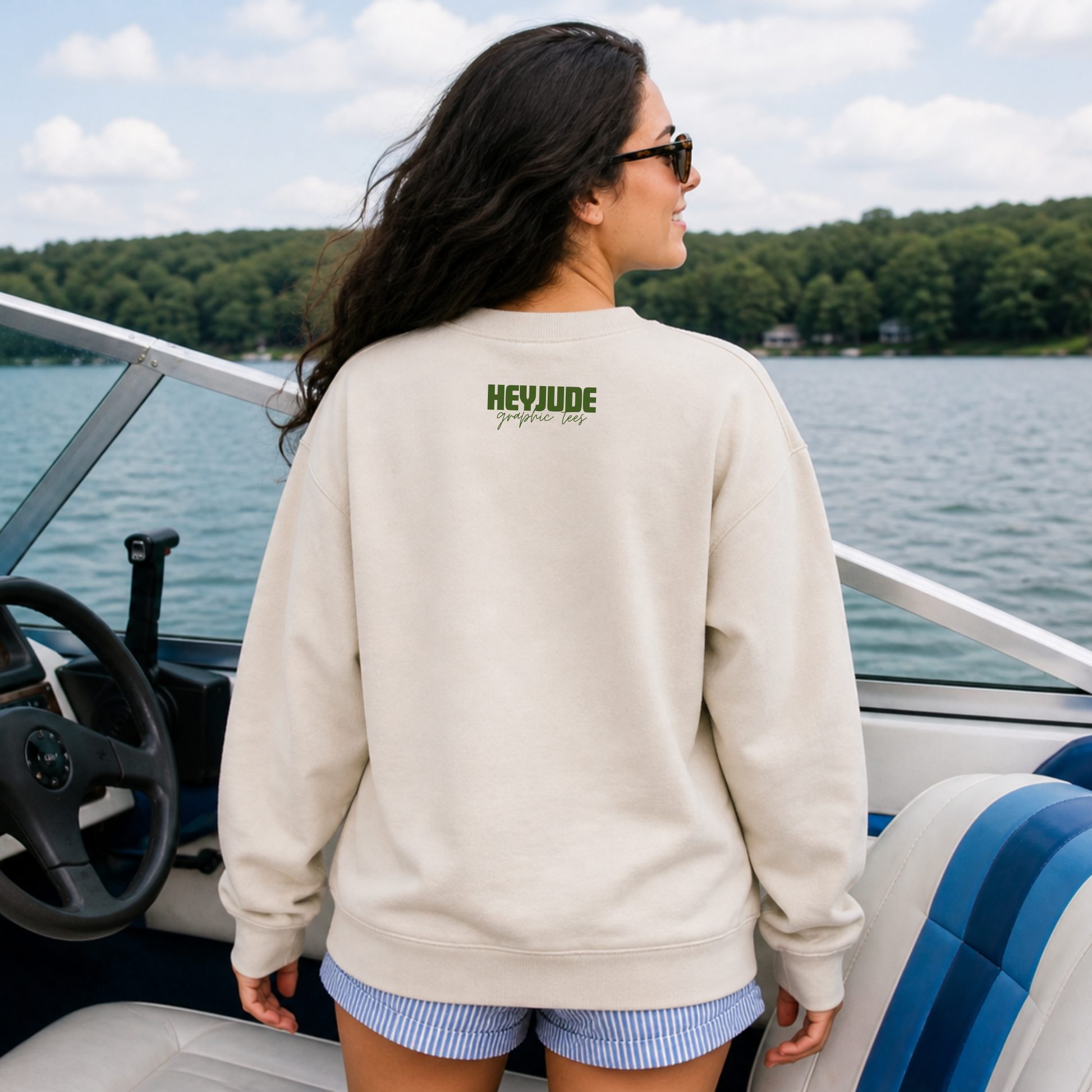 Woman wearing a beige sweatshirt with 'HEYJUDE' logo on a boat by a lake.