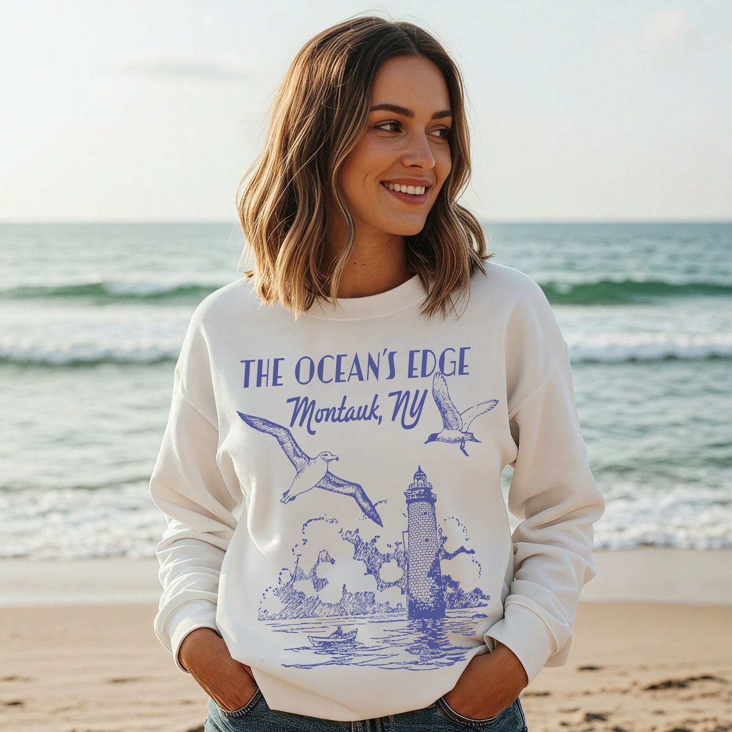 "Smiling woman at the beach wearing white Montauk ‘The Ocean’s Edge’ coastal graphic sweatshirt."